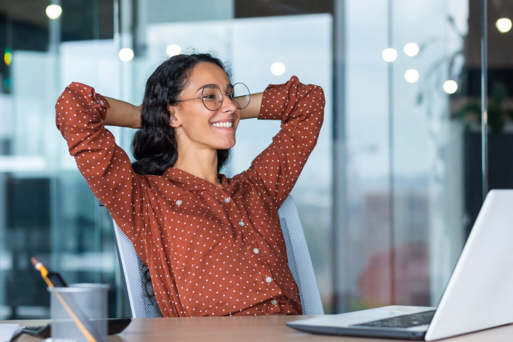young professional woman at desk with her hands behind her head smiling